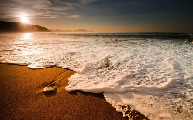 El sol saliendo al mar detrás de las montañas y la espuma de las olas golpeando la playa 2K fondo de pantalla
