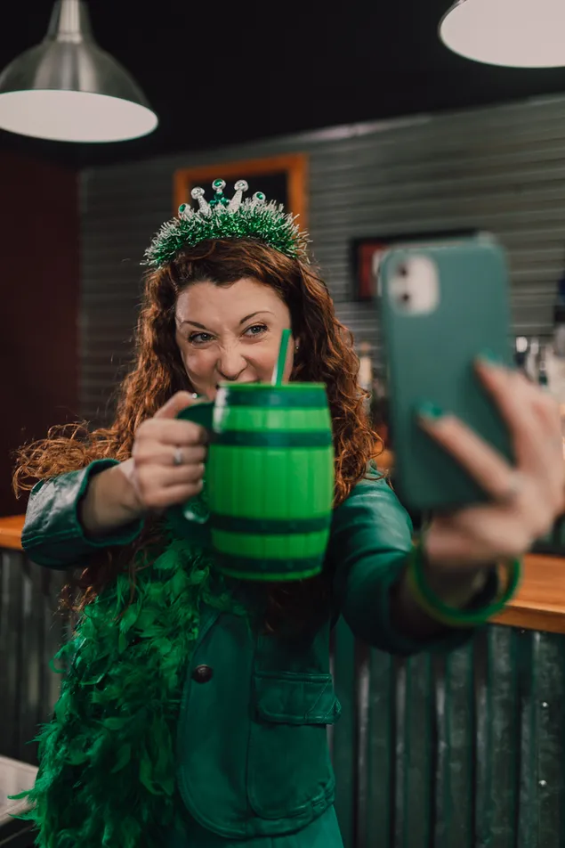 Muat turun Wanita yang meminum minuman Hari Saint Patrick dan mengambil swafoto dengan pakaian hijau