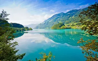 Tiny houses in a small village at the foot of high mountains and trees, forests and high hills reflected in the lake water 2K wallpaper