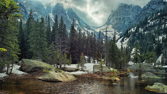 Bäume und ruhiger fließender Fluss zwischen schneebedeckten Bergen und Hügeln 2K Hintergrundbild