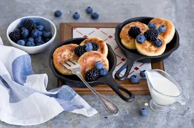 dve panvice palaciniek s modrymi a ciernymi bobulami a mliekom v podnose stiahnut