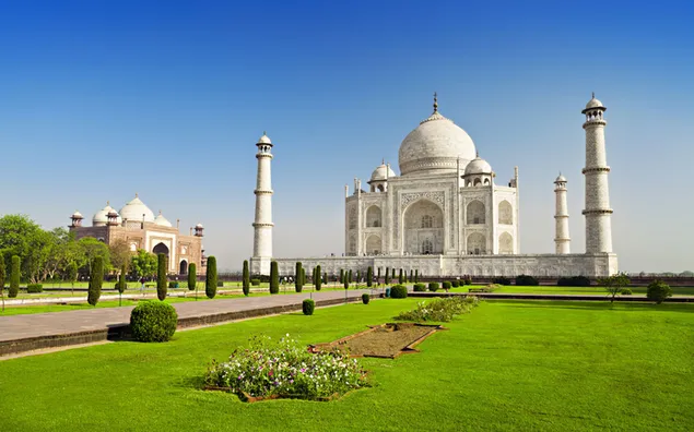 Uitsig op Taj Mahal, 'n unieke argitektoniese werk in wit, met manjifieke groen plante en helder lug aflaai