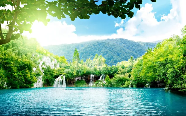 Air terjun yang mengalir melalui pegunungan dan pepohonan dengan sinar matahari yang cerah di balik dedaunan dan awan unduhan