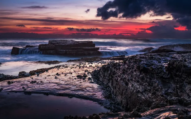 Wellen von Meerwasser zwischen Felsen im Rot des Sonnenuntergangs hinter dunklen Wolken