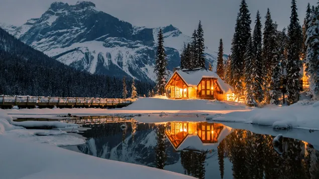 Wooden house with lights on the snowy foothills and reflection of trees in the lake
