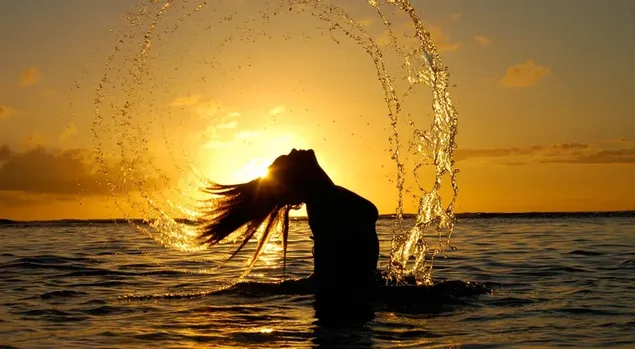夕日を背景にビーチで髪で水を投げる女の子